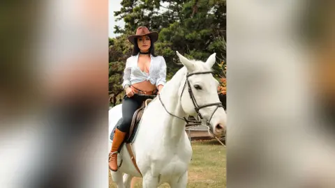 FlorenciaCopper sits upright on a white horse wearing a white button-down shirt and black leggings or tight pants.