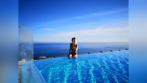 Blonde woman in a bikini relaxing on the edge of a luxury infinity pool overlooking the blue ocean at sunset.