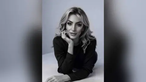 JessycaPage poses with her hand on her chin, looking at the camera with long blonde hair and a black top.