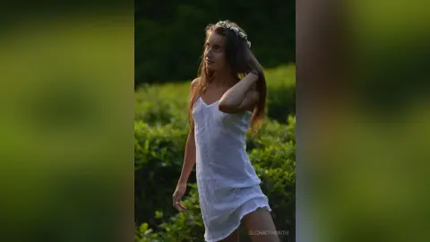 Dreamy portrait of a young woman in a white summer dress and flower crown standing in a lush green field.
