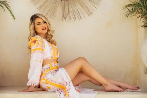 Blonde woman in a floral white and yellow summer dress relaxing on a sunlit terrace in a bohemian setting.