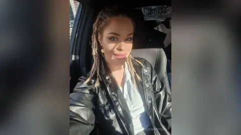 Smiling woman with braided hair wearing a black leather jacket sitting in a car on a sunny day.