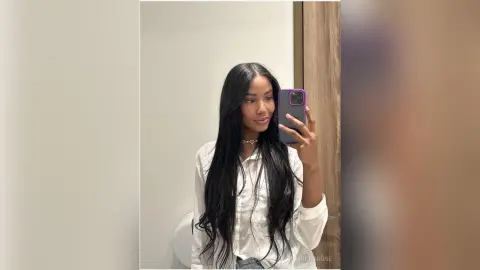 Young woman with long dark hair in a white button-down shirt taking a casual mirror selfie in a bright room.