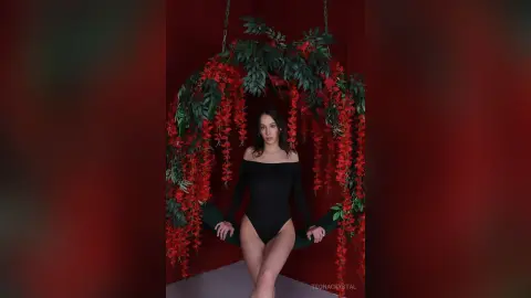 Elegant woman in a black bodysuit sitting within a lush red floral swing against a deep red background.
