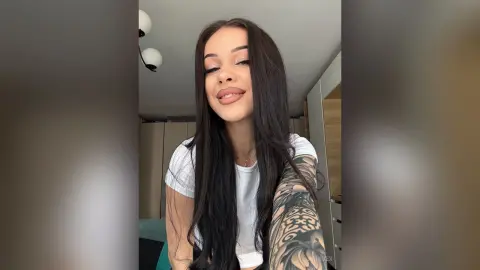 AriaVex smiles while wearing a white t-shirt, showing off her long dark hair and large arm tattoo.