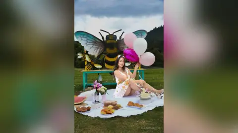 Woman in white floral dress enjoying a summer picnic with cake and fruit near a giant bee decoration.