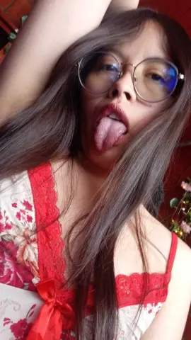Young woman with long hair and glasses wearing a floral red and white lace camisole in a cozy indoor setting.