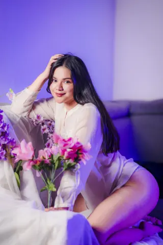 Smiling young woman in a white long-sleeve shirt sitting amidst pink flowers in soft purple lighting.
