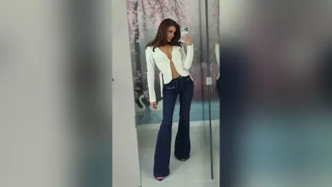 AnaMathers poses in a white open-front bolero top and dark blue flared jeans while taking a mirror selfie.