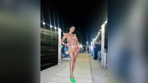 NicoleBella stands on a wooden walkway wearing a tight, long-sleeved, multi-colored patterned mini dress and bright green heels.