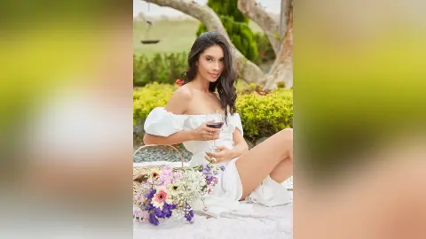 LucyMartin sits outdoors in a white off-the-shoulder dress, holding a glass of red wine next to a bouquet of flowers.