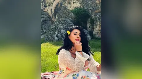 Beautiful woman in a white lace cardigan and floral dress sitting on green grass near rocky mountains.