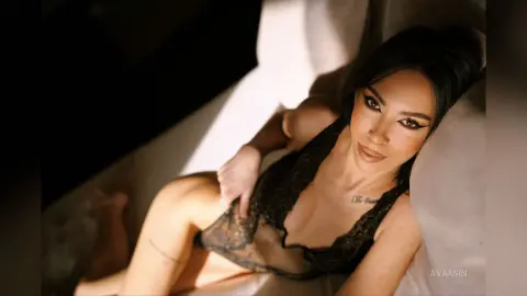 AvaaSin lies back in a black lace bodysuit, showing her cleavage and dark hair against a white background.