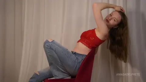 Redheaded woman in a red lace crop top and blue jeans sitting on a red chair against white curtains.