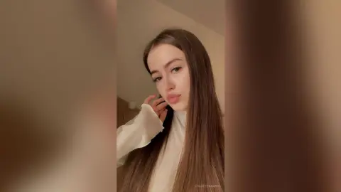 Young woman with long brown hair wearing a white turtleneck sweater in a soft, minimalist indoor setting.