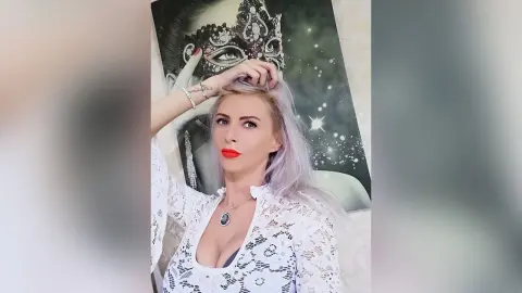 Elegant woman with silver hair in a white lace top posing in front of a large masquerade mask artwork.