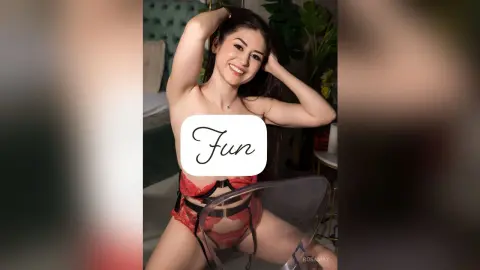 RosaMay smiles while posing naked with her hands behind her head, wearing red lace lingerie and black strap details.