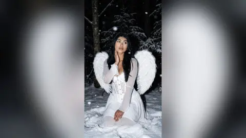 Ethereal woman in a white angel costume with large wings sitting in a snowy winter forest at night.