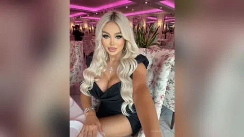 AlexaEvans poses in a black dress with a deep neckline, showing off her long blonde hair and silver cross necklace in a pink-lit room.