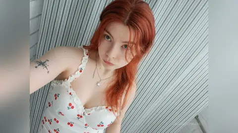 Redheaded woman in a white floral sundress posing indoors against striped curtains with a soft mood.