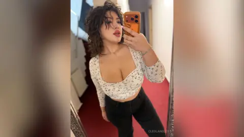 Young woman with curly hair in a floral white crop top taking a mirror selfie in a cozy room.