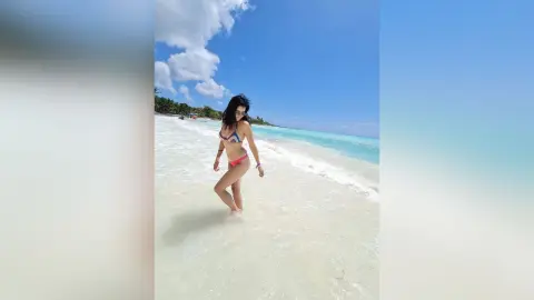 SandraMidnight walks through shallow turquoise ocean water on a white sand beach wearing a pink bikini.