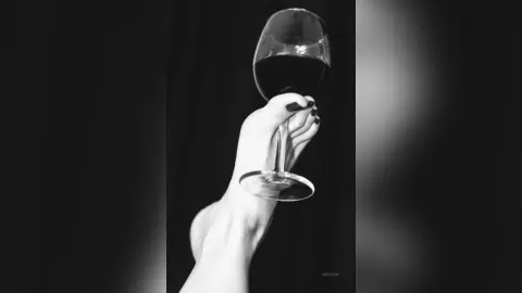 AryaSin holds a glass of red wine against her bare foot, showing her dark painted toenails against the glass stem.