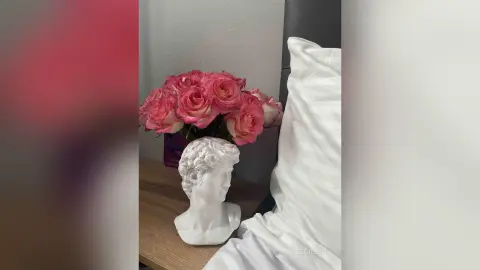 Pink roses in a vase next to a white classical bust sculpture on a wooden bedside table in a bright room.