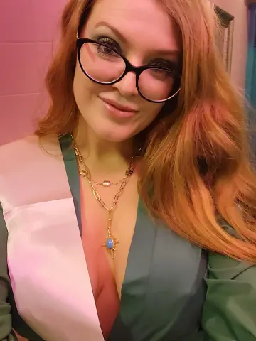 Anna_OG smiles wearing black glasses, gold necklaces, and a green top with a white sash over her chest.