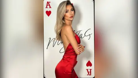 Elegant woman in a red satin dress posing against a giant Ace of Hearts playing card in a minimalist studio.