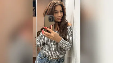 Brunette woman in a grey houndstooth long-sleeve top taking a stylish mirror selfie in a modern setting.
