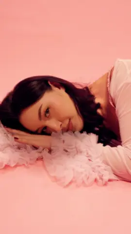 Smiling young woman in a white ruffled dress lying on a pink background for a dreamy aesthetic portrait.