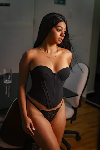 Young woman in black corset and lace lingerie posing in a dimly lit room with a moody atmosphere.