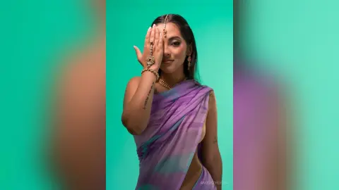 Elegant woman in a purple silk saree and gold jewelry posing against a teal background.