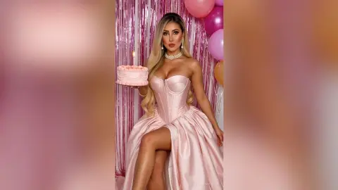 Glamorous woman in a pink satin corset gown holding a birthday cake against a festive pink fringe backdrop.