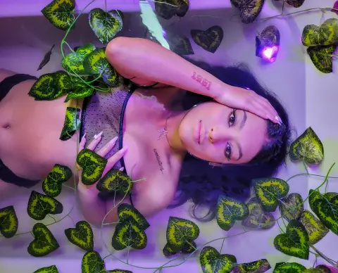 Young woman in black bikini relaxing in a bathtub filled with green heart-shaped leaves under purple lighting.