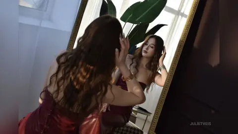 Woman in a red satin dress looking in a gold mirror in a bright, modern room.