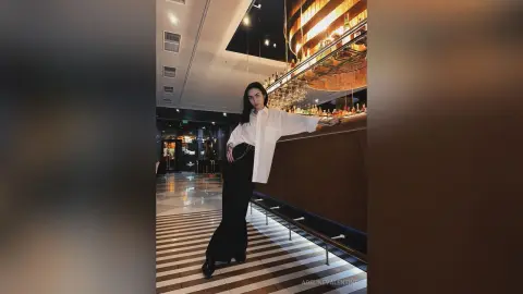 Stylish woman in a white button-down shirt and black trousers standing in a modern, moody lounge bar.