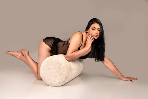 Curvy woman in black sheer lingerie leaning on a white textured pouf against a neutral studio background.