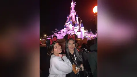 LucyMartin smiles wearing red sequined mouse ears and a grey sweatshirt against a lit purple castle at night.