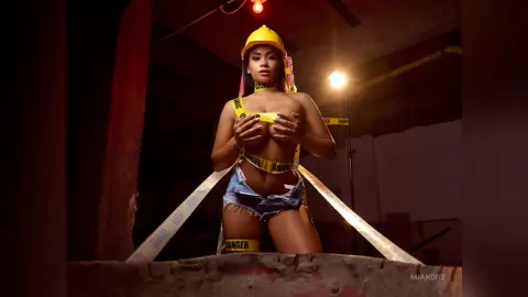 MiaKorz wears a yellow hard hat and yellow danger tape across her bare breasts and denim shorts in a dark construction site setting.