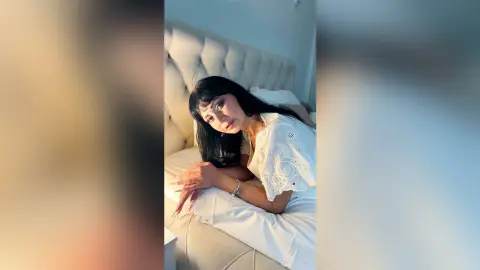 Woman in white eyelet blouse relaxing on a bed in a bright, cozy bedroom setting.