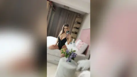 Young woman in a black bodysuit taking a mirror selfie in a bright, modern bedroom.