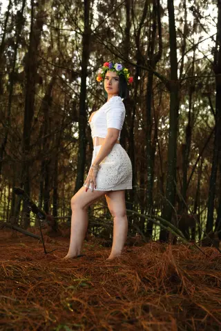 Woman in white summer outfit and flower crown standing in a dreamy, sunlit pine forest.
