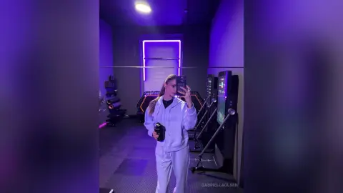 Young woman in a white tracksuit taking a mirror selfie in a modern gym with purple neon lighting.