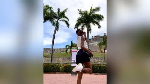 Stylish Black woman in a white top and black shorts posing near palm trees at a sunny tropical historic site.
