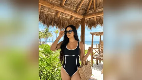 Woman in a black one-piece swimsuit and sunglasses relaxing under a tropical straw hut by the beach.