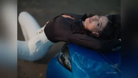 Stylish woman in a black blouse and white jeans relaxing on a blue car in a moody outdoor setting.