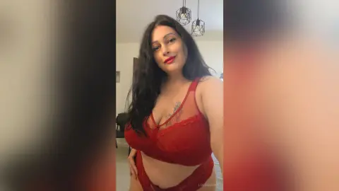 Woman in red lace lingerie posing confidently in a modern bedroom setting.
