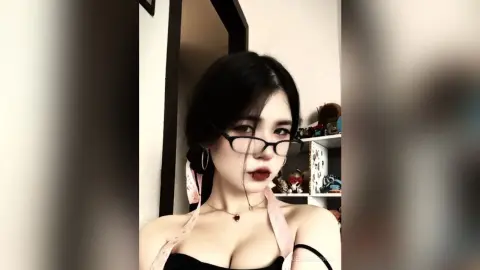 DeaneMerrih wears black glasses and a black top with pink straps, showing her cleavage and dark hair in a close-up shot.